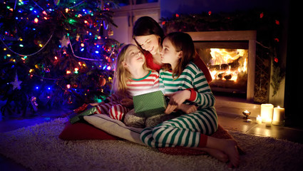 Happy young mother and her two small daughters opening a magical Christmas gift by a fireplace in a cozy dark living room on Christmas eve.