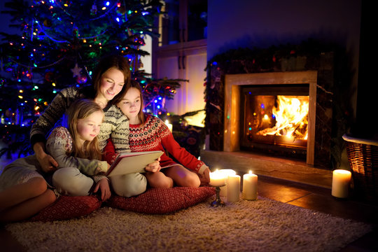 Mother and her two cute young daughters using a tablet pc at home by a fireplace in warm and cozy living room on Christmas eve.