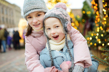 Fototapeta premium Two adorable sisters having a good time together on traditional Christmas fair in Riga, Latvia. Children enjoying sweets, candies and gingerbread on Xmas market.