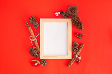 Centered wooden frame with white copy space for text on red background with autumn decorations: pine cones, cinnamon sticks, leaves and branches, with vintage christmas decorations