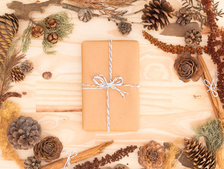 Fototapeta na wymiar Centered kraft paper rustic gift box isolated on a wooden table background with autumn decorations: pine cones, cinnamon sticks, leaves and branches / For weddings, fall, winter and holidays