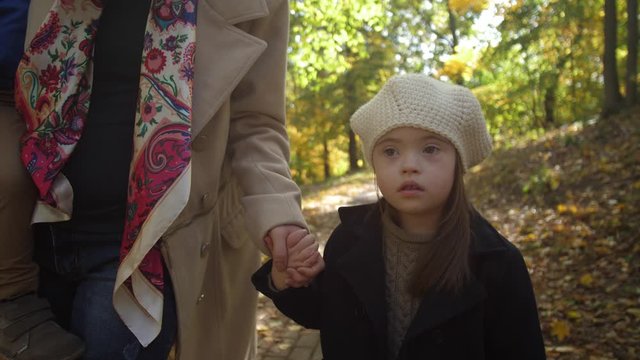 Serious little girl with down syndrome walking hand with mother through autumn park. Midsection of loving mom holding toddler son on her arms and leading special needs daughter by hand in nature