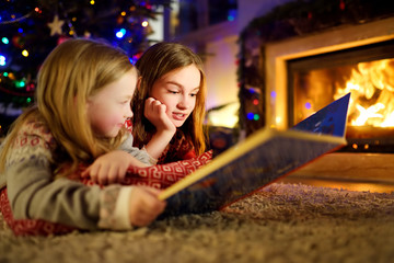 Happy young sisters reading a story book together by a fireplace in a cozy dark living room on...