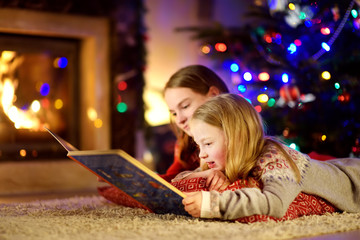 Happy young sisters reading a story book together by a fireplace in a cozy dark living room on...
