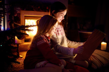 Happy young sisters reading a story book together by a fireplace in a cozy dark living room on...