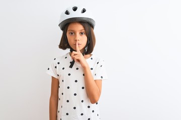 Beautiful child girl wearing security bike helmet standing over isolated white background asking to be quiet with finger on lips. Silence and secret concept.