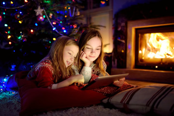 Two cute young sisters using a tablet pc at home by a fireplace in warm and cozy living room on Christmas eve.