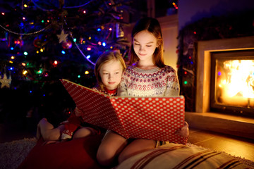 Happy young sisters reading a story book together by a fireplace in a cozy dark living room on Christmas eve. Celebrating Xmas at home.