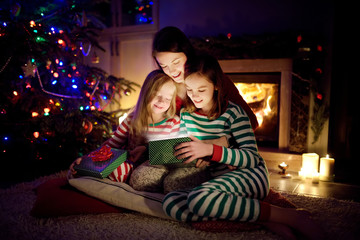 Fototapeta na wymiar Happy young mother and her two small daughters opening a magical Christmas gift by a fireplace in a cozy dark living room on Christmas eve.
