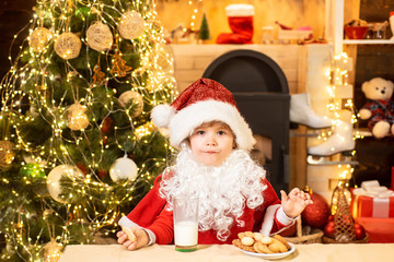 Fototapeta na wymiar Happy Santa Claus - cute boy child eating a cookie and drinking glass of milk at home Christmas interior. Santa - funny child picking cookie.