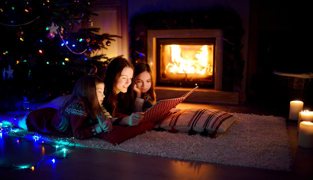 Happy young mother and her daughters reading a story book together by a fireplace in a cozy dark living room on Christmas eve. Celebrating Xmas at home.