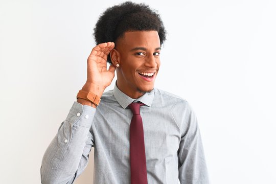 Young african american businessman wearing tie standing over isolated white background smiling with hand over ear listening an hearing to rumor or gossip. Deafness concept.
