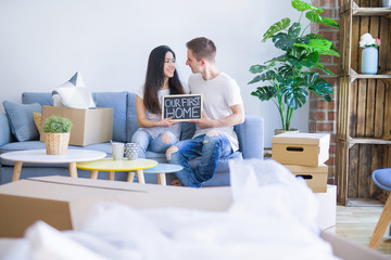 Young beautiful couple sitting on the sofa holding blackboard with message at new home around cardboard boxes