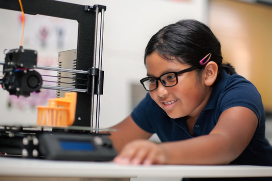 A happy young girl wearing glasses and watching a 3d printer finish the 3d model she created.