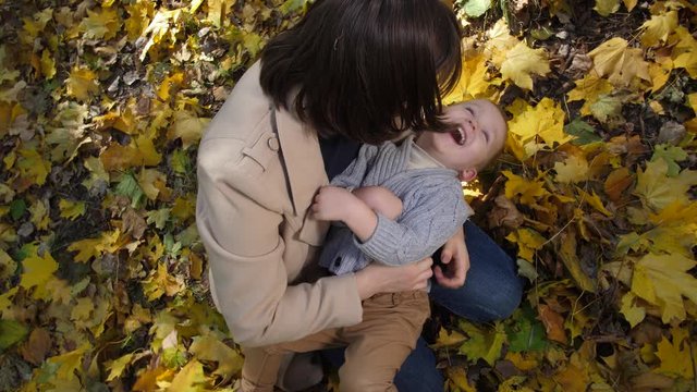 Above view of cute little boy joyfully laughing from tickling while lying on knees of playful mother sitting among yellow autumn leaves in park. Happy family enjoying fun during joint outdoor leisure