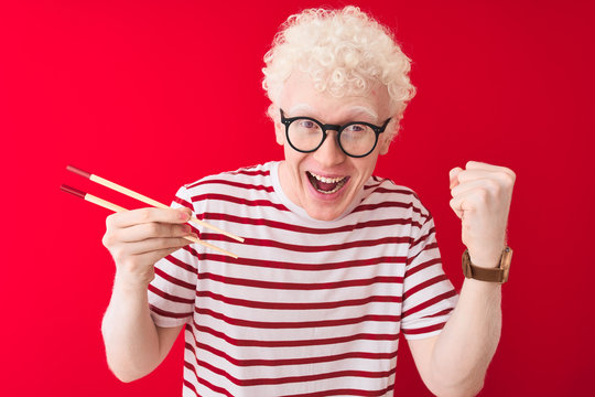 Young albino blond man holding chopsticks standing over isolated white background very happy and excited, winner expression celebrating victory screaming with big smile and raised hands