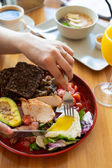 Woman eating breakfast with knife and fork in a cafe: avocado and nuts, rye toasts, vegetables, fried bacon, chicken and poached egg