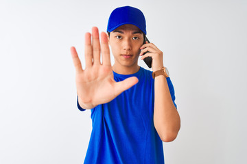 Chinese deliveryman wearing cap talking on the smartphone over isolated white background with open hand doing stop sign with serious and confident expression, defense gesture