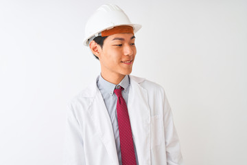 Chinese architect man wearing coat and helmet standing over isolated white background looking away to side with smile on face, natural expression. Laughing confident.