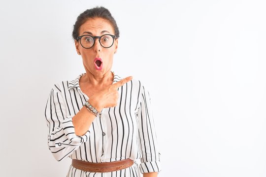 Middle age businesswoman wearing striped dress and glasses over isolated white background Surprised pointing with finger to the side, open mouth amazed expression.