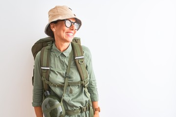Middle age hiker woman wearing backpack canteen hat glasses over isolated white background looking away to side with smile on face, natural expression. Laughing confident.