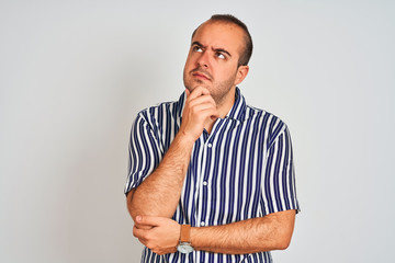 Young man wearing blue striped shirt standing over isolated white background with hand on chin thinking about question, pensive expression. Smiling with thoughtful face. Doubt concept.