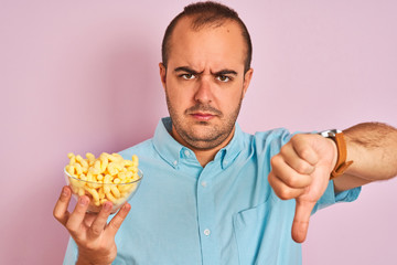 Young man holding bowl with extruded corn standing over isolated pink background with angry face, negative sign showing dislike with thumbs down, rejection concept