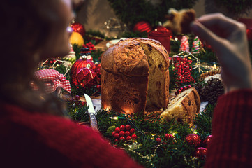 Young woman eating a piece of panettone, in the background a table of Christmas ingredients accompanied by hot chocolate, orange, cookies, cinnamon, pine. - obrazy, fototapety, plakaty