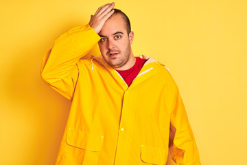 Young man wearing rain coat standing over isolated yellow background surprised with hand on head for mistake, remember error. Forgot, bad memory concept.