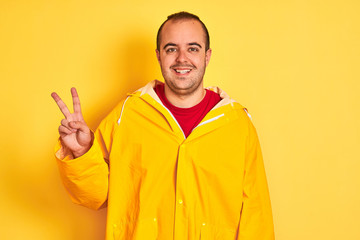 Naklejka premium Young man wearing rain coat standing over isolated yellow background smiling with happy face winking at the camera doing victory sign. Number two.