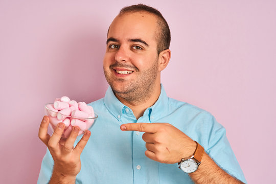 Young man holding bowl with marshmallows standing over isolated pink background very happy pointing with hand and finger