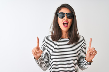 Chinese woman wearing striped t-shirt and sunglasses standing over isolated white background amazed and surprised looking up and pointing with fingers and raised arms.