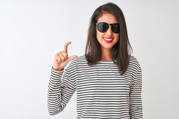 Naklejka premium Chinese woman wearing striped t-shirt and sunglasses standing over isolated white background smiling and confident gesturing with hand doing small size sign with fingers looking and the camera