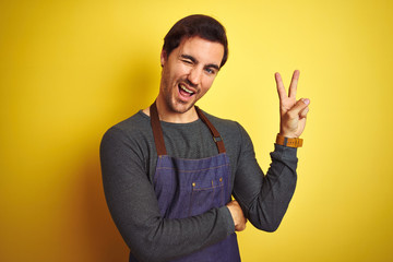 Young handsome shopkeeper man wearing apron standing over isolated yellow background smiling with happy face winking at the camera doing victory sign. Number two.