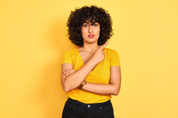 Young arab woman with curly hair wearing t-shirt standing over isolated yellow background Pointing with hand finger to the side showing advertisement, serious and calm face