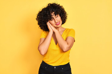 Young arab woman with curly hair wearing t-shirt standing over isolated yellow background sleeping tired dreaming and posing with hands together while smiling with closed eyes.