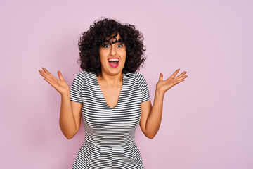Young arab woman with curly hair wearing striped dress over isolated pink background celebrating crazy and amazed for success with arms raised and open eyes screaming excited. Winner concept