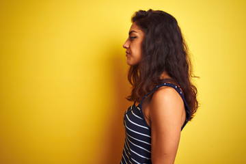 Young beautiful woman wearing striped t-shirt standing over isolated yellow background looking to side, relax profile pose with natural face and confident smile.