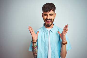 Young man with tattoo wearing blue shirt standing over isolated white background celebrating mad...