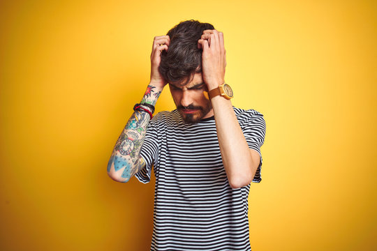 Young man with tattoo wearing striped t-shirt standing over isolated yellow background suffering from headache desperate and stressed because pain and migraine. Hands on head.