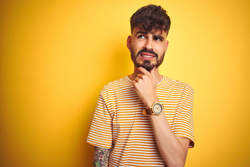 Young man with tattoo wearing striped t-shirt standing over isolated yellow background Thinking worried about a question, concerned and nervous with hand on chin