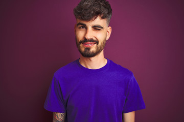 Obraz premium Young man with tattoo wearing t-shirt standing over isolated purple background winking looking at the camera with sexy expression, cheerful and happy face.