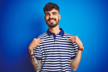 Young man with tattoo wearing striped polo standing over isolated blue background looking confident with smile on face, pointing oneself with fingers proud and happy.