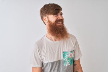 Young redhead irish man wearing t-shirt standing over isolated white background looking away to side with smile on face, natural expression. Laughing confident.