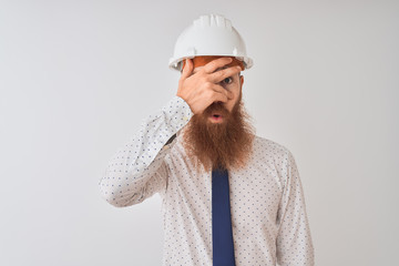 Young redhead irish architect man wearing security helmet over isolated white background peeking in shock covering face and eyes with hand, looking through fingers with embarrassed expression.