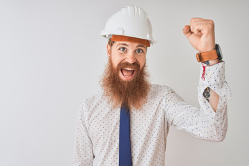 Young redhead irish architect man wearing security helmet over isolated white background angry and mad raising fist frustrated and furious while shouting with anger. Rage and aggressive concept.