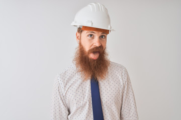 Young redhead irish architect man wearing security helmet over isolated white background sticking tongue out happy with funny expression. Emotion concept.