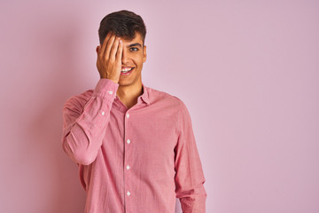 Young indian man wearing elegant shirt standing over isolated pink background covering one eye with hand, confident smile on face and surprise emotion.