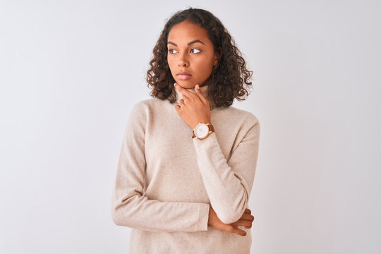 Young brazilian woman wearing turtleneck sweater standing over isolated white background looking confident at the camera with smile with crossed arms and hand raised on chin. Thinking positive.