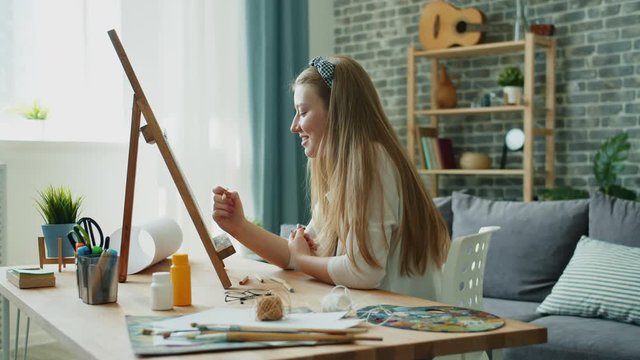Joyful student attractive blond girl is painting picture at home at easel, paintbrush and palette visible on table. Fine arts, hobby and creative youth concept.